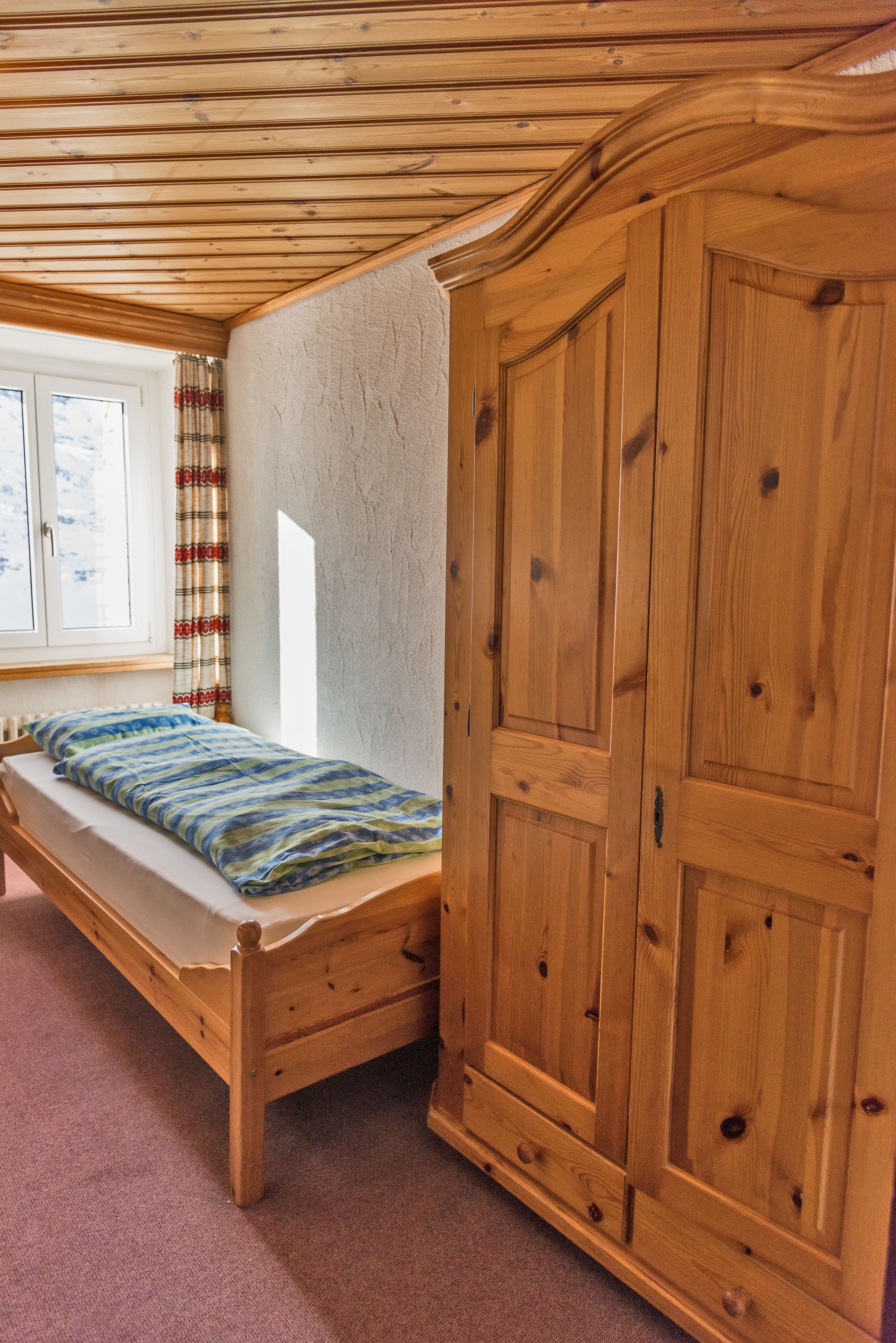A wooden bed with a mattress and pillows, a wooden wardrobe, and a window with curtains.