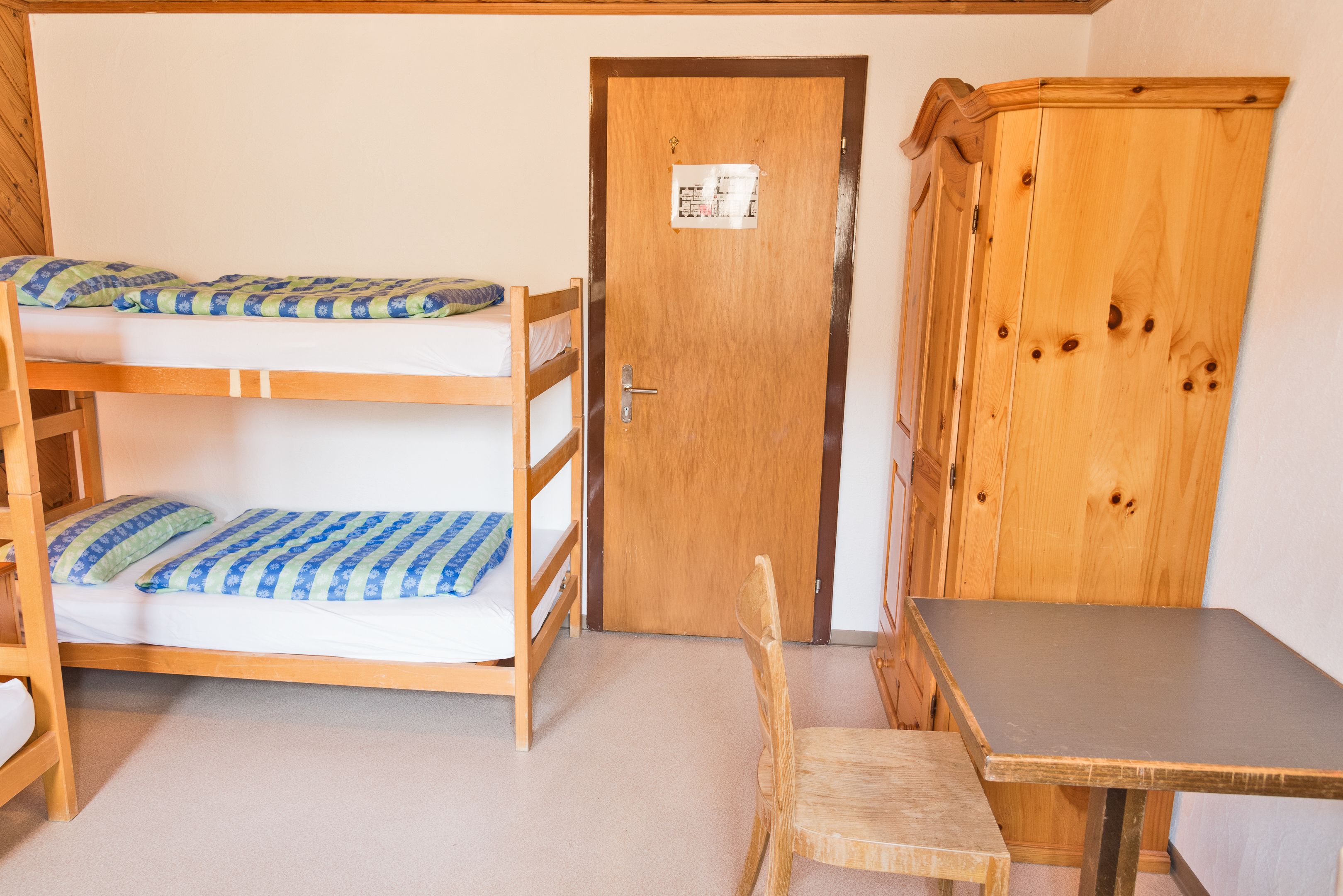 A bunk bed with a wooden headboard, a wooden cabinet, and a small table in a room with white walls and carpet.