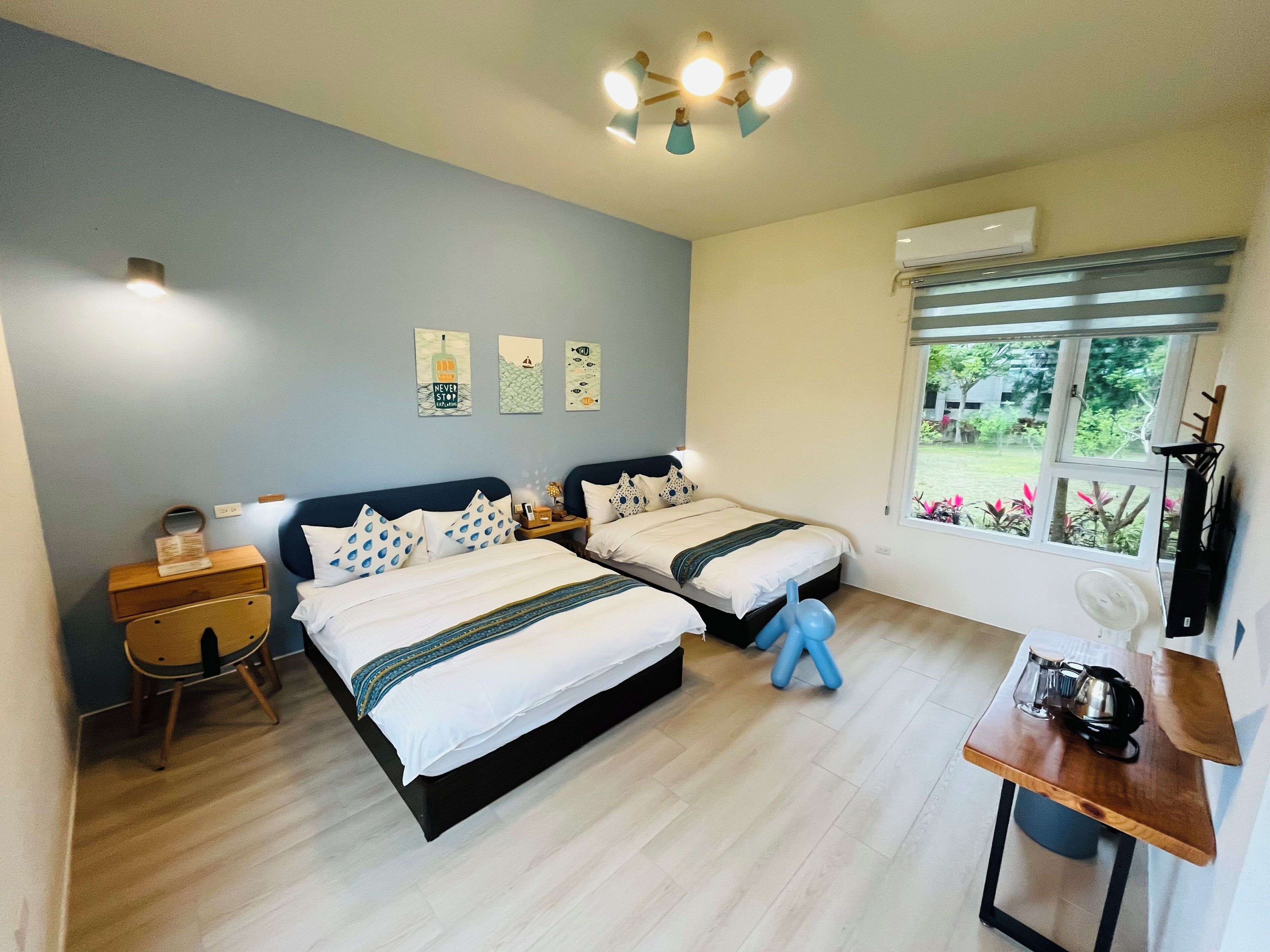 Quarto com duas camas, mesa de cabeceira de madeira, escrivaninha e janela com vista para vegetação.