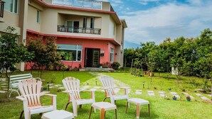 Un bâtiment moderne à la façade rouge et beige, doté d’un balcon et d’un jardin bien entretenu, agrémenté de chaises et de plantes en pot.
