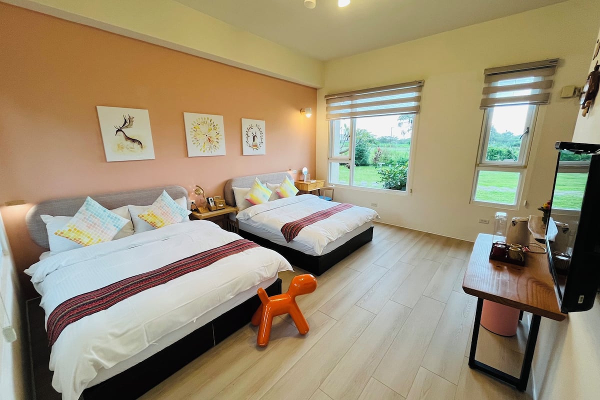 A bedroom with two beds, a small table, a TV, and a window with a view of greenery.
