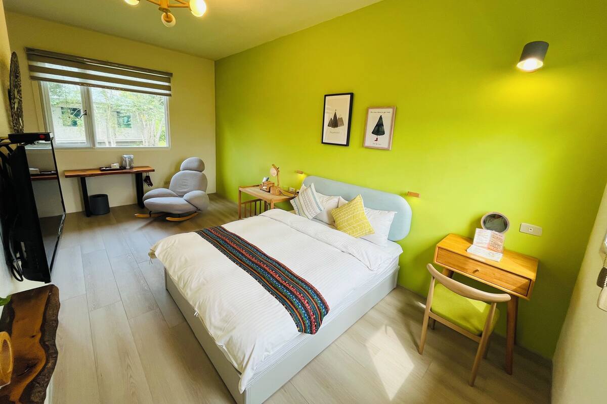 A bedroom with a bed, a desk, a chair, a bedside table, and a window.