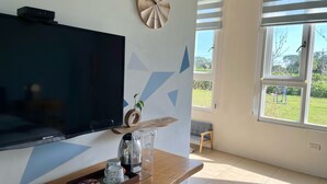Un salon moderne doté d’un téléviseur à écran plat, d’une table basse en bois sur laquelle se trouve un plateau avec divers objets, d’une horloge décorative et d’une fenêtre donnant sur l’extérieur.