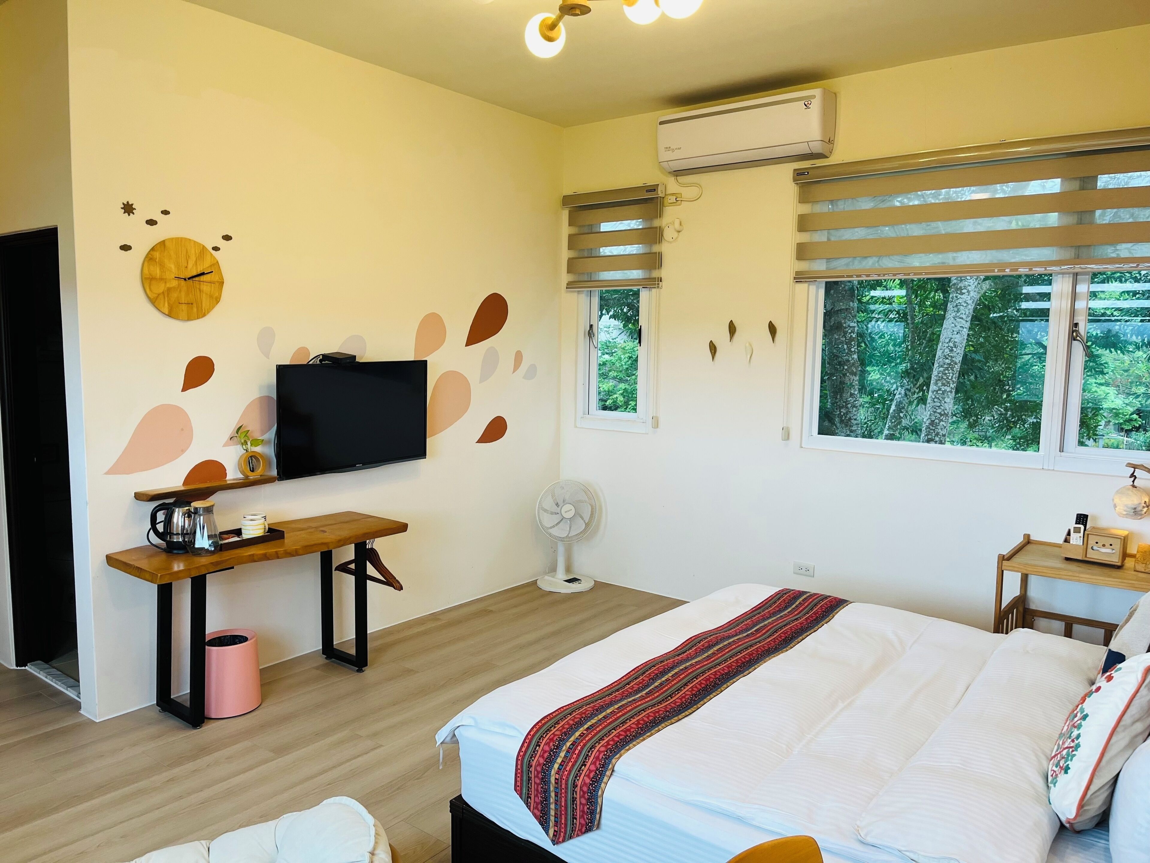 A bedroom with a bed, a TV, a desk, a fan, and a window with blinds.