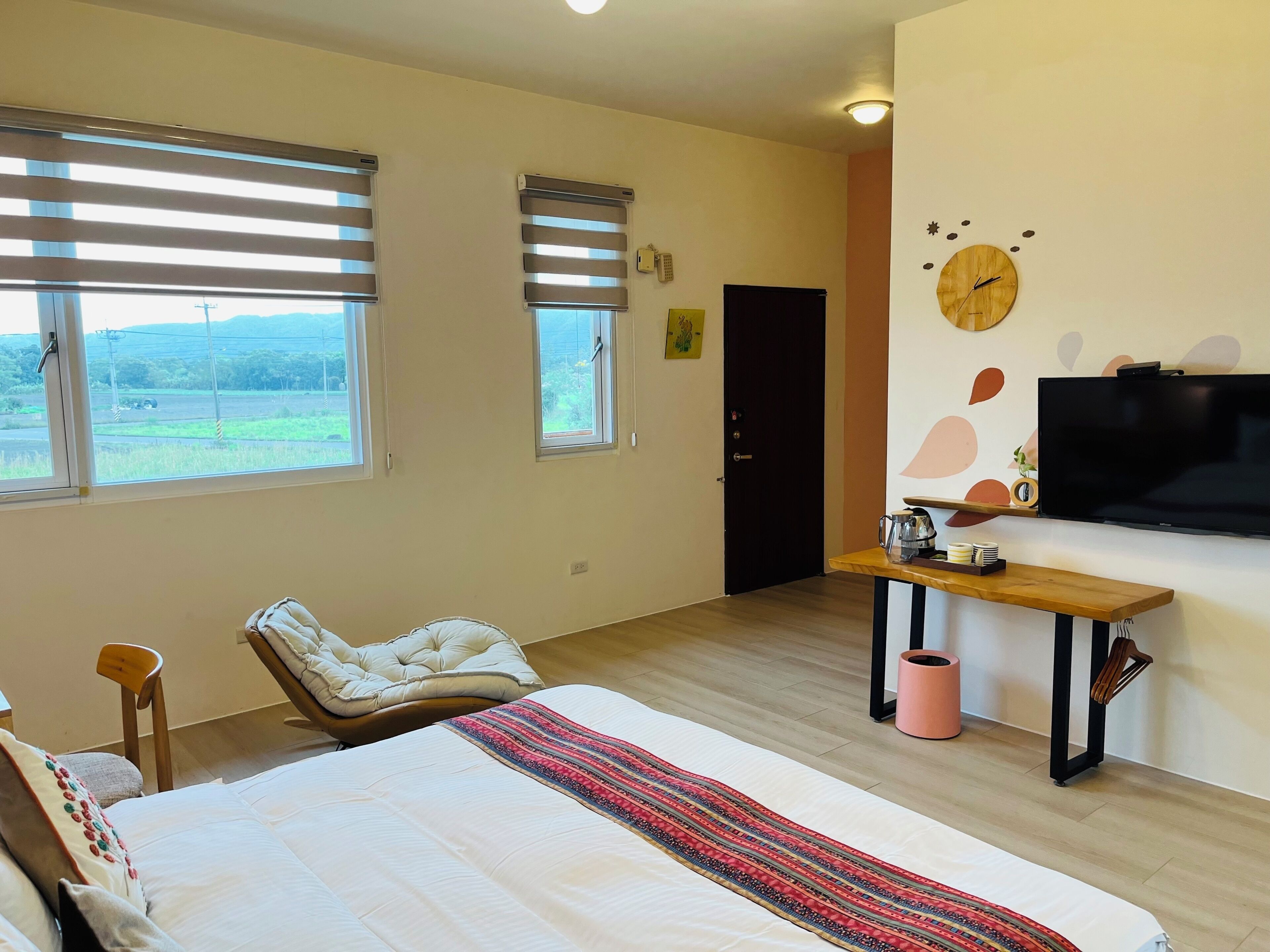 A bedroom with a bed, a chair, a desk, a TV, and windows with blinds.