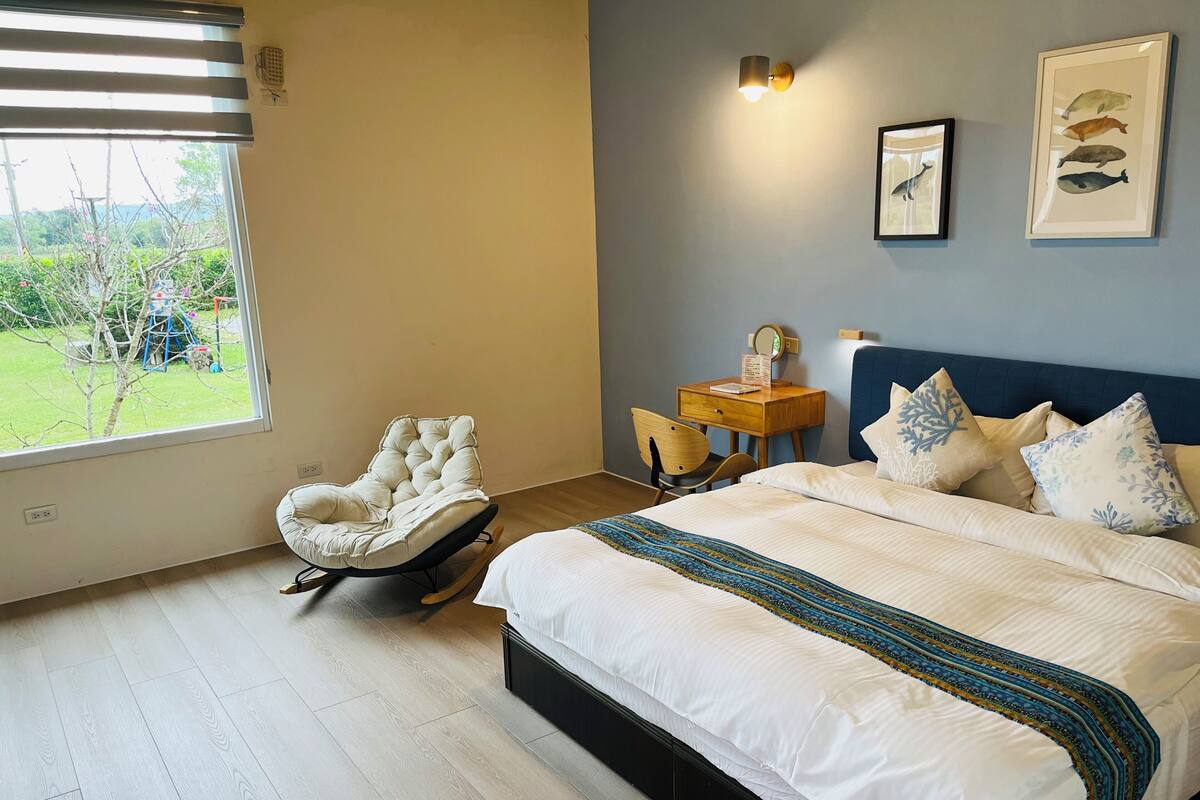 A bedroom with a bed, a rocking chair, a desk with a chair, a window, and wall art.