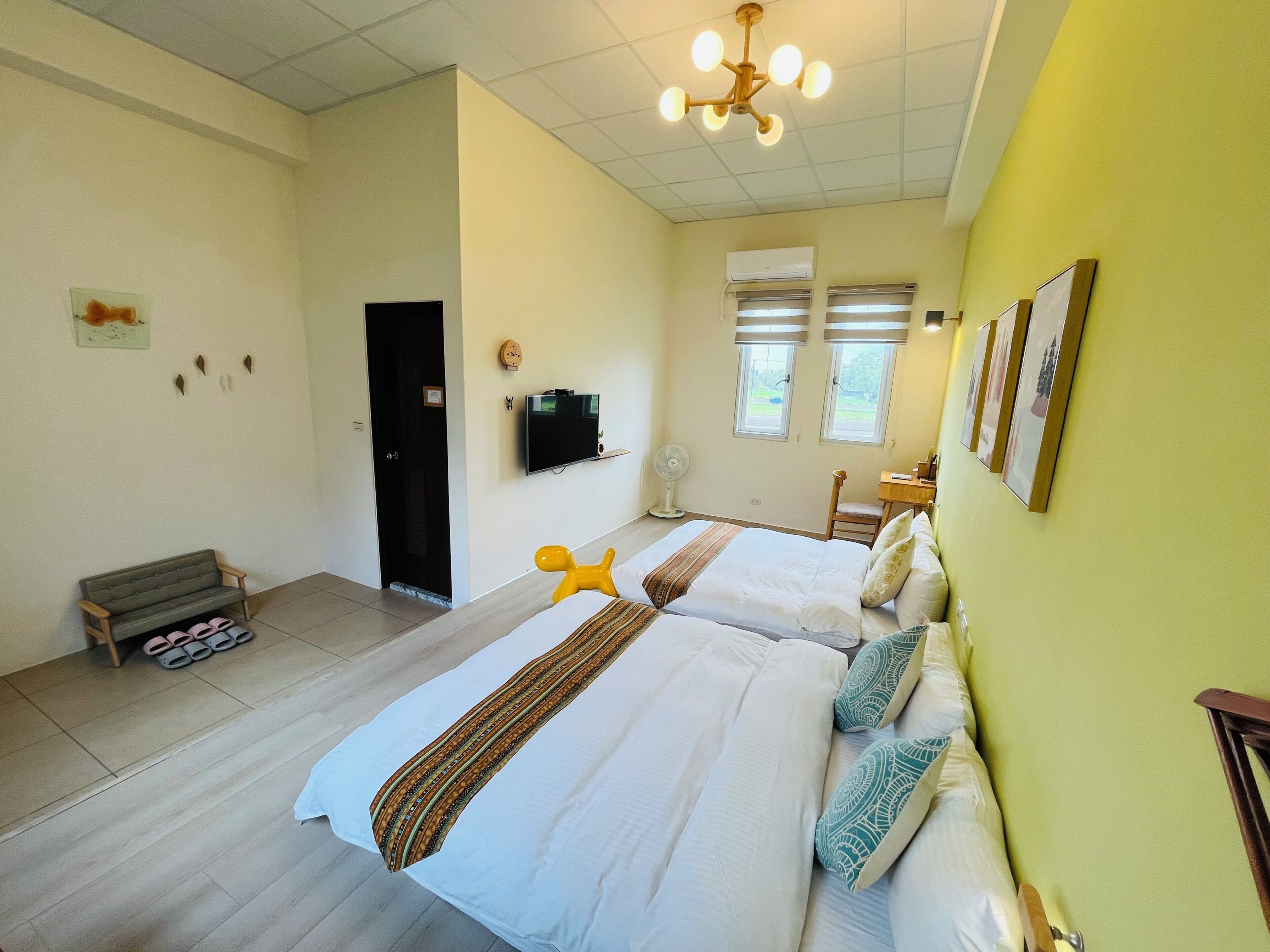Quarto de hotel com duas camas, uma escrivaninha, uma cadeira, uma televisão e um ventilador.