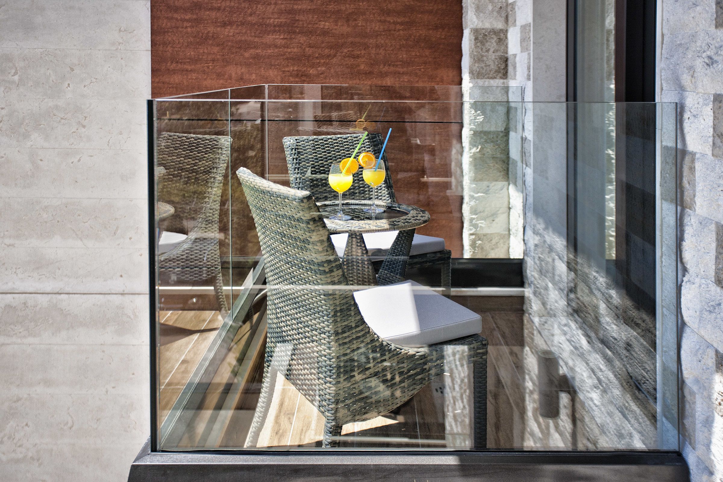 A glass-enclosed balcony with wicker chairs and a small table, featuring a glass of orange juice and a cocktail with a straw.
