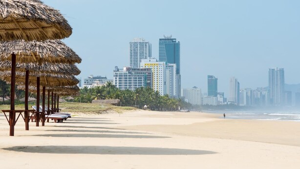 A beach with thatched umbrellas and lounge chairs, a city skyline with high-rise buildings, and a clear sky. - New Sky Hotel (Da Nang)