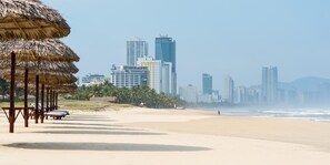 A beach with thatched umbrellas and lounge chairs, a city skyline with high-rise buildings, and a clear sky. - New Sky Hotel (Da Nang)