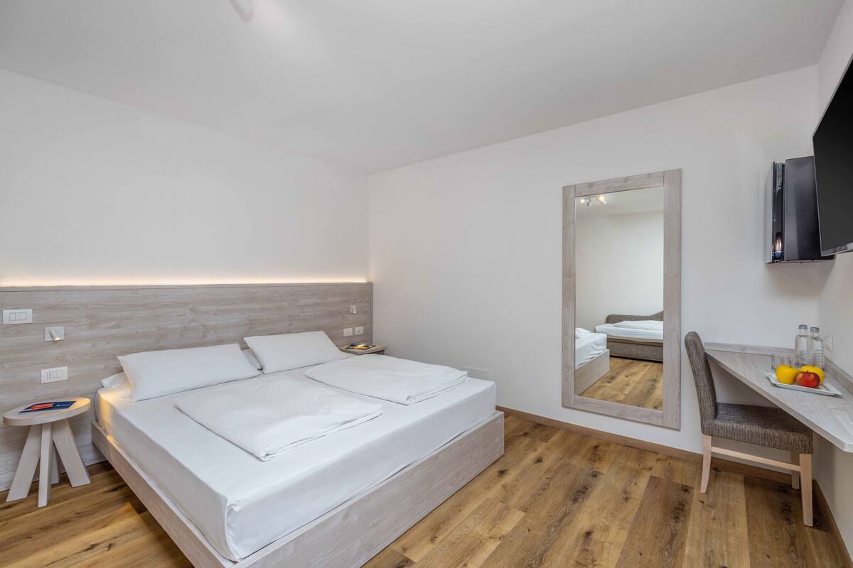 A modern hotel room with a large bed, a bedside table, a mirror, a desk with a chair, and a flat-screen TV.