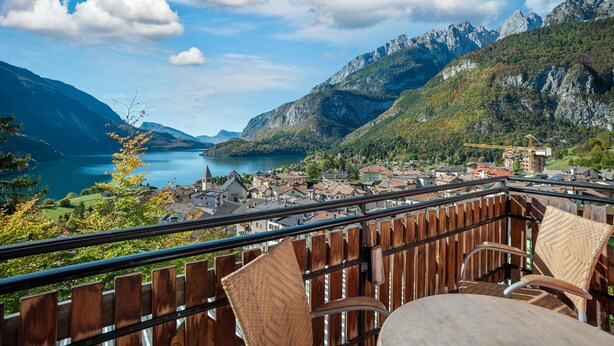 A balcony with wicker chairs and a table overlooking a lake and mountains. - Hotel Londra (Molveno)