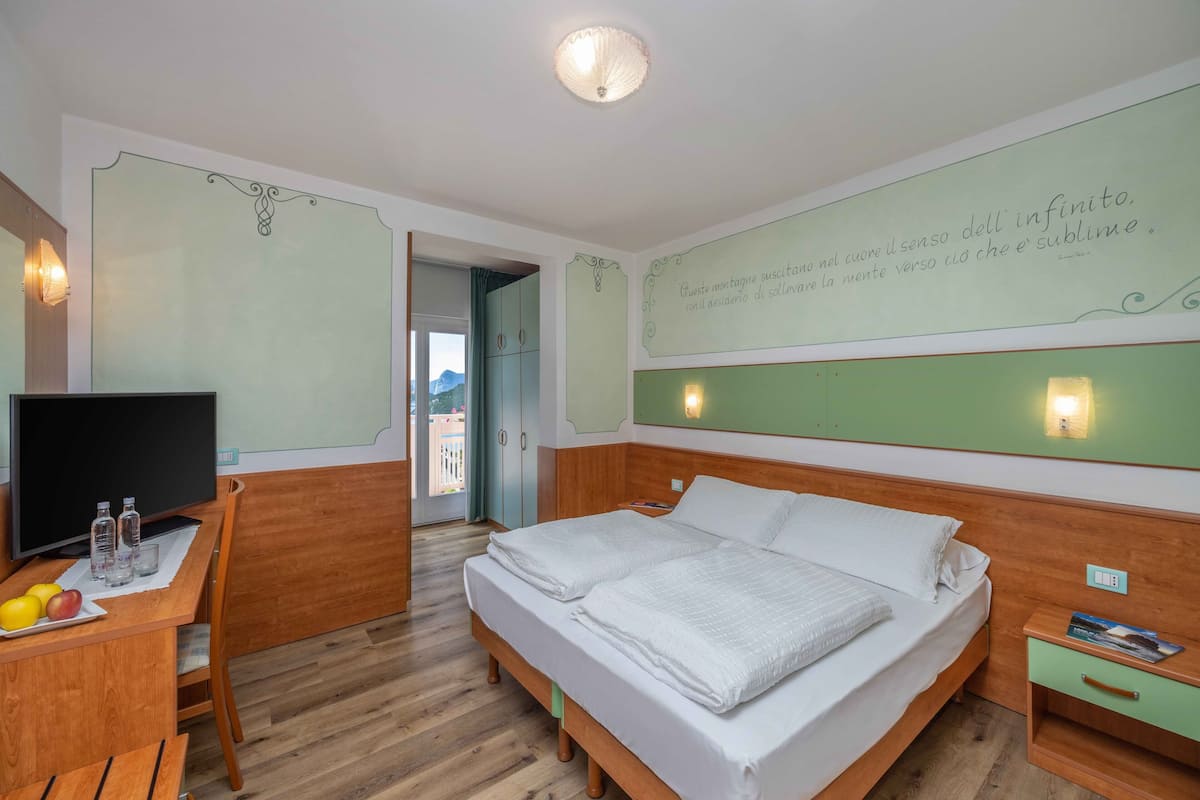 A hotel room with a bed, a desk with a TV, a chair, a nightstand, and a wall with a quote.