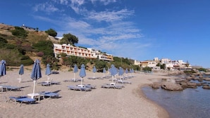 A beach with sun loungers and umbrellas, a building with a white facade, and rocky terrain. - Creta Mare (Agios Vasileios)