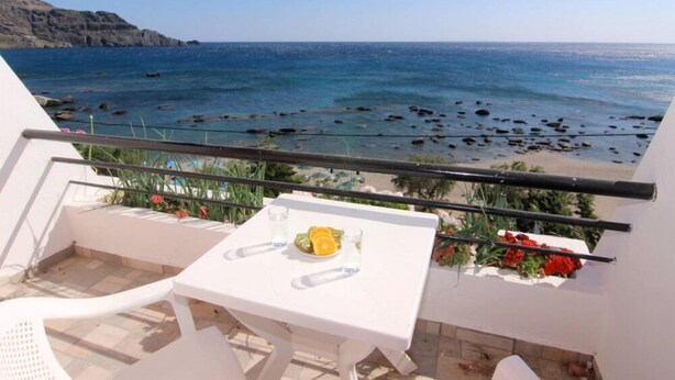 A balcony with a table and chairs, overlooking a rocky beach and the sea. - Creta Mare (Agios Vasileios)