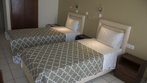 A hotel room with two beds, each with a patterned bedspread, bedside tables, and a small wooden nightstand. - Creta Mare (Agios Vasileios)