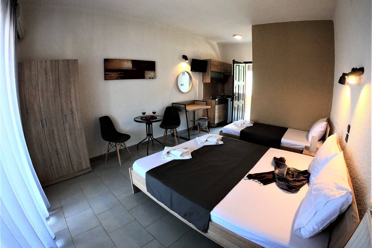 Ein Hotelzimmer mit zwei Betten, einem Schreibtisch, einem Stuhl, einem kleinen Tisch mit zwei Weingläsern und einer Küchenzeile.