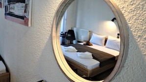 Un miroir rond reflète un lit bien fait, avec des draps blancs et une tête de lit en bois, une table de chevet avec une lampe, et un cadre photo fixé au mur.