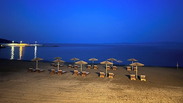 Une plage bordée de rangées de transats et de parasols, une jetée en arrière-plan et une eau calme qui reflète les lumières.