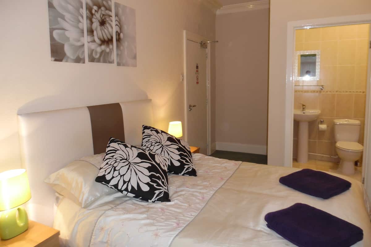 A bedroom with a bed, two pillows, a nightstand with a lamp, a bathroom with a sink and toilet, and a shower area.
