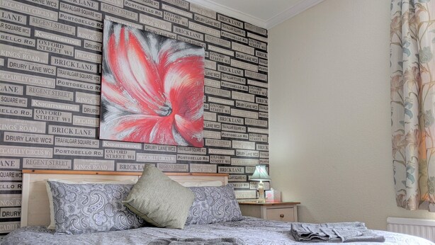 A neatly made bed with patterned bedding, a large floral artwork on the wall, and a nightstand with a lamp and a folded towel. - Kirk Lea Guest House (Halifax)