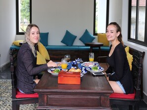 Dos personas sentadas en una mesa de madera con platos de comida, un vaso de zumo de naranja y una tetera. En el fondo, hay un sofá azul con cojines amarillos.