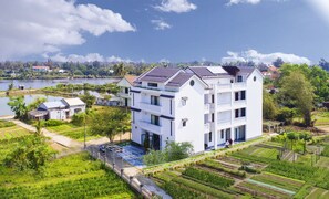 Un edificio blanco de varios pisos con balcones, rodeado de vegetación exuberante y una masa de agua.