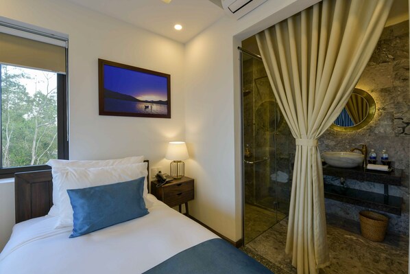 A bedroom with a bed, a nightstand, a lamp, a picture on the wall, a bathroom with a sink and shower curtain.