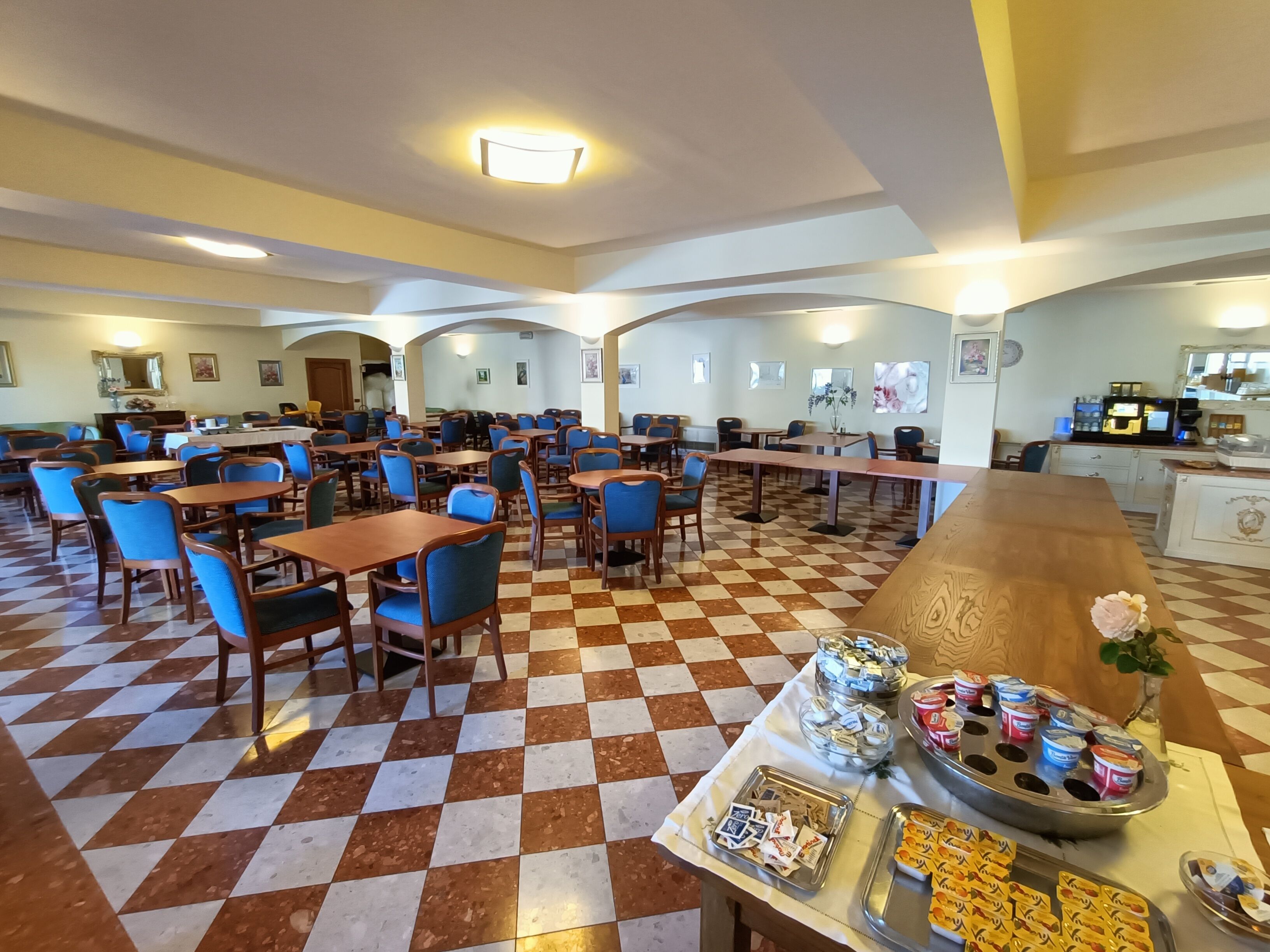 Ein großzügiger Essbereich mit kariertem Boden, mehreren Tischen und Stühlen sowie einem Buffet mit verschiedenen Speisen.