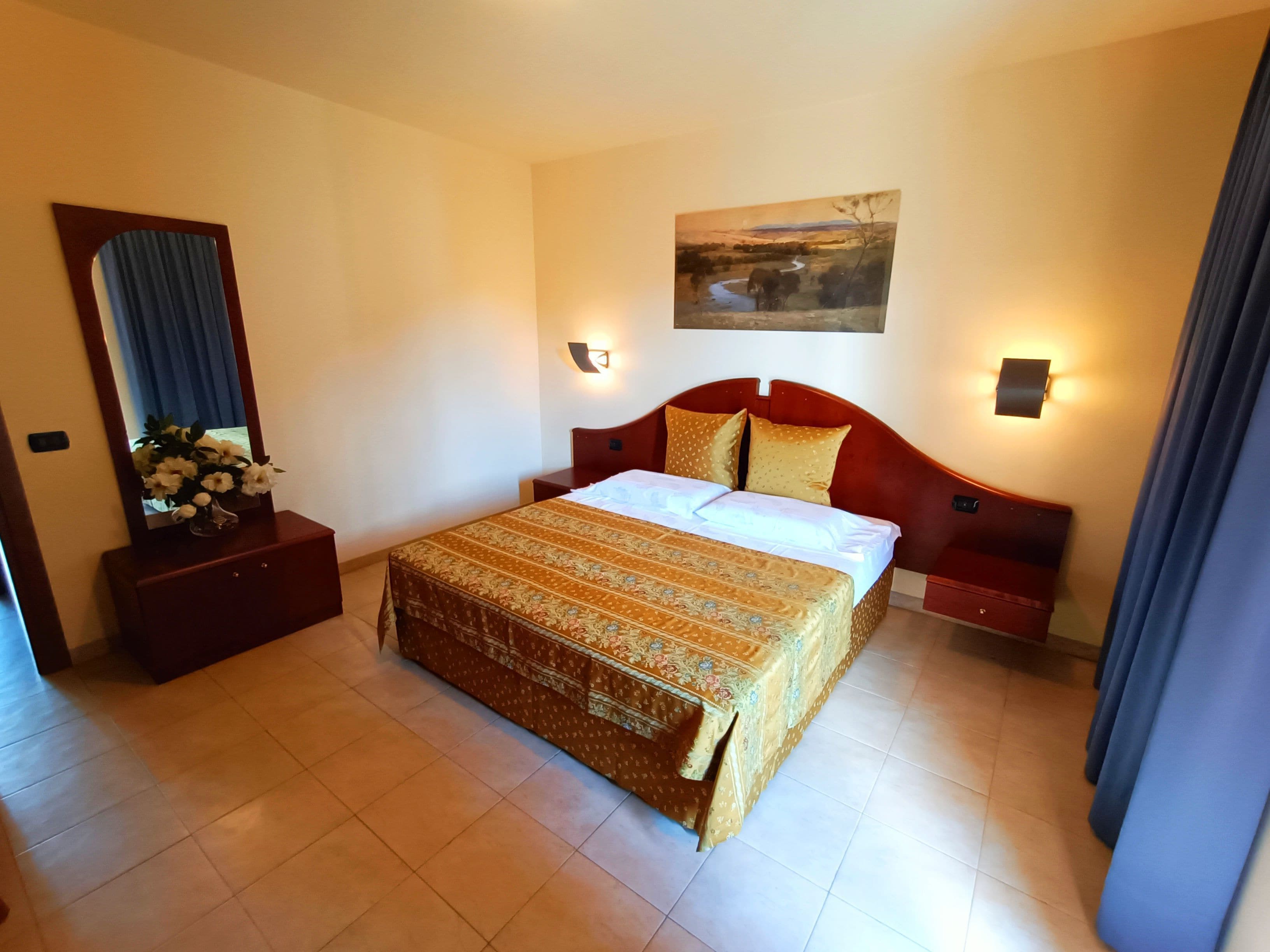 A hotel room with a large bed, a nightstand with a vase of flowers, a wall-mounted painting, and a mirror.