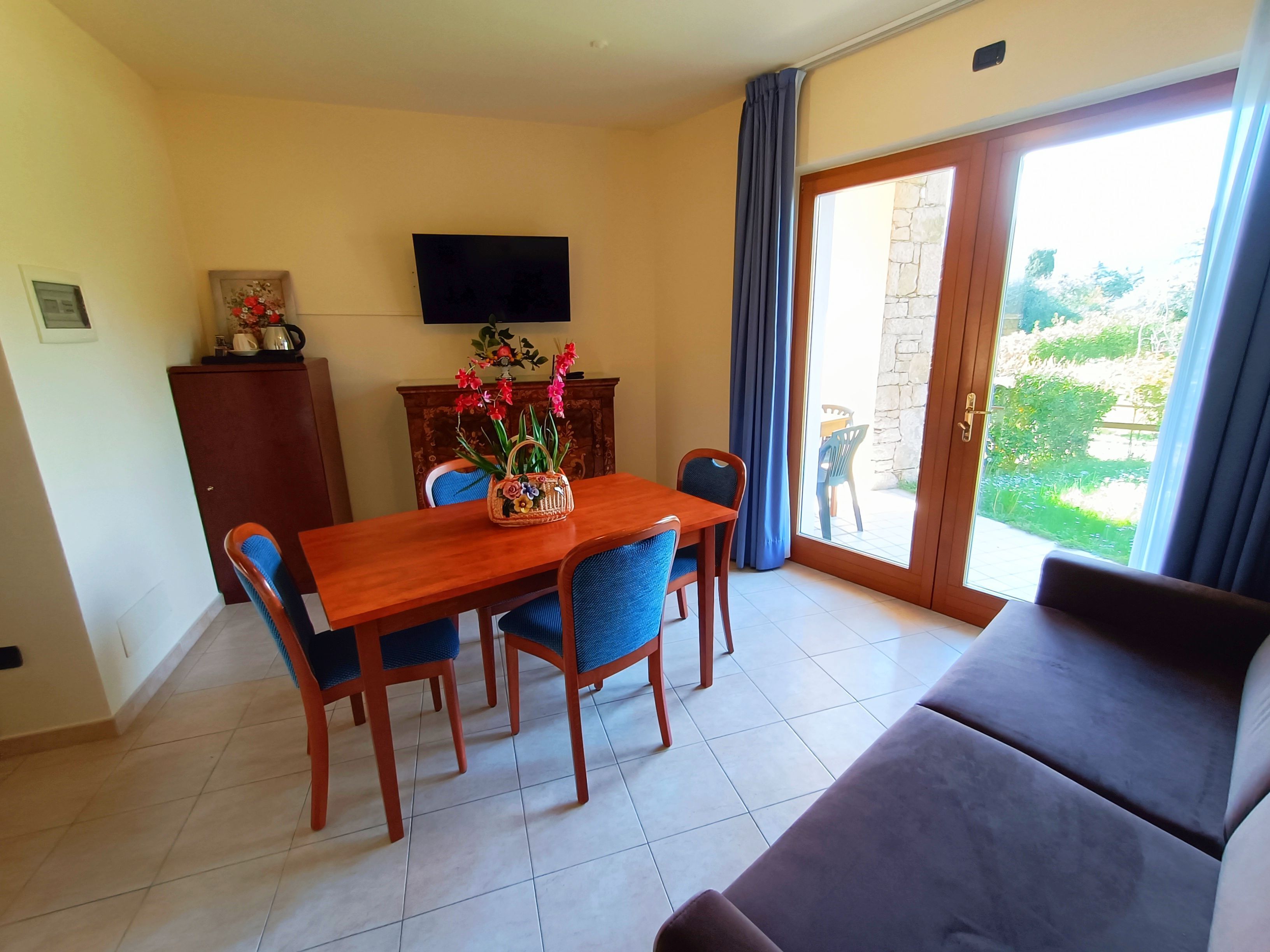 Un comedor con mesa de madera y sillas azules, un sofá, un televisor en la pared y vistas al exterior a través de un ventanal.