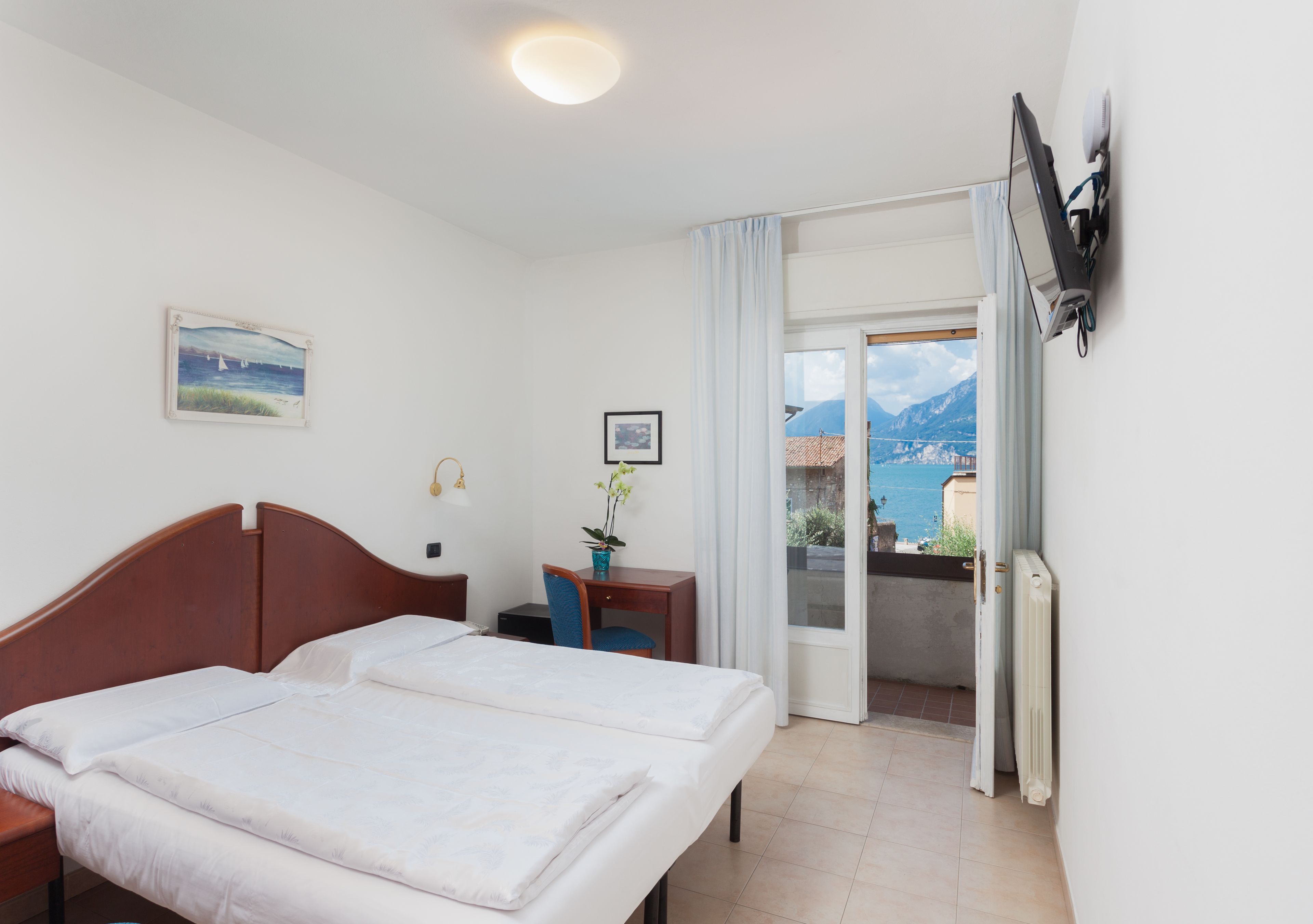 A hotel room with two beds, a desk, a chair, a potted plant, a painting, and a view of the sea and mountains through a sliding door.
