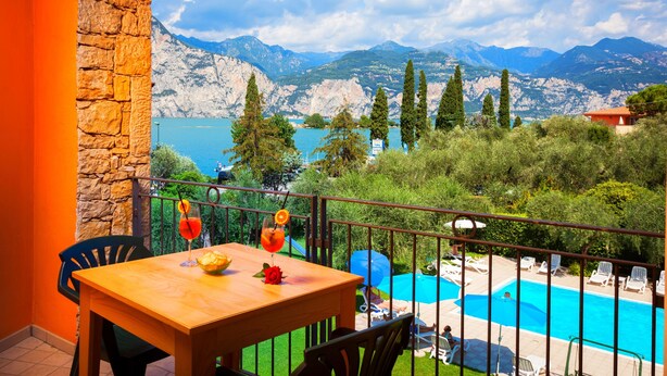 A balcony with a wooden table, chairs, and a view of a swimming pool and mountains. - Villa Isabella Hotel & Residence (Brenzone sul Garda)