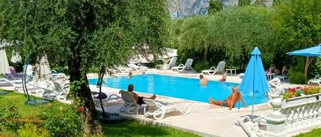 A pool area with lounge chairs and umbrellas, surrounded by greenery and mountains in the background.