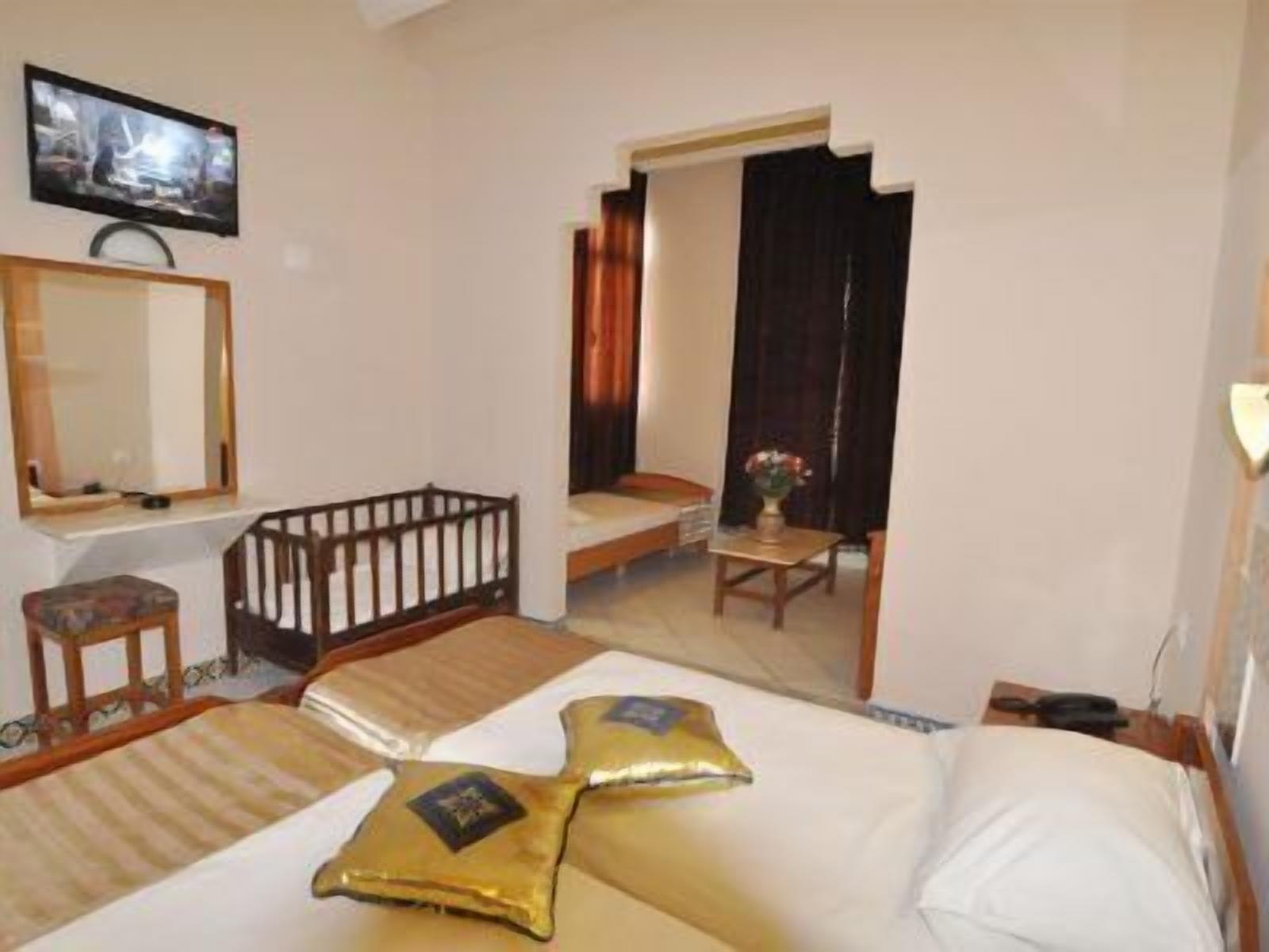A hotel room with a bed, a small table with a vase, a chair, a crib, and a television on the wall.