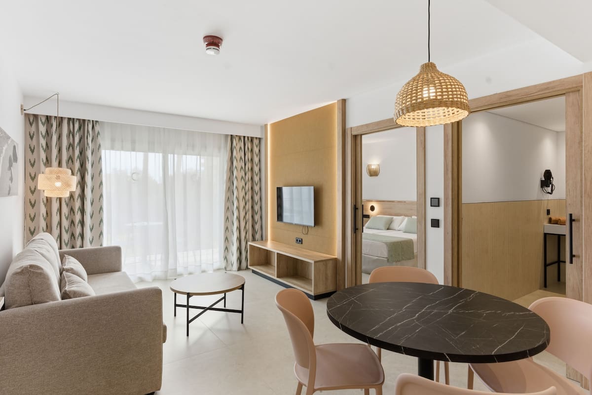 A modern hotel room with a sofa, dining table, chairs, a TV, and a large window with curtains.