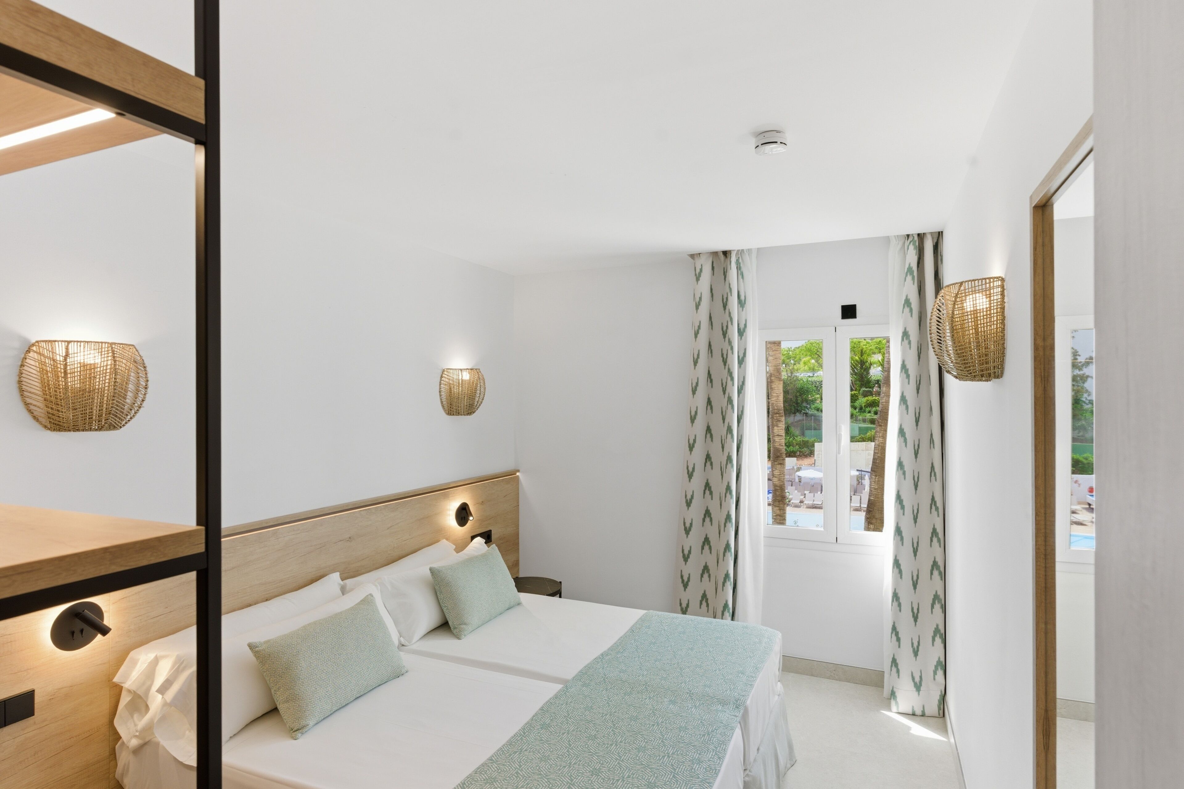 Habitación de hotel con cama, lámparas de noche, ventana con cortinas y vistas al exterior.