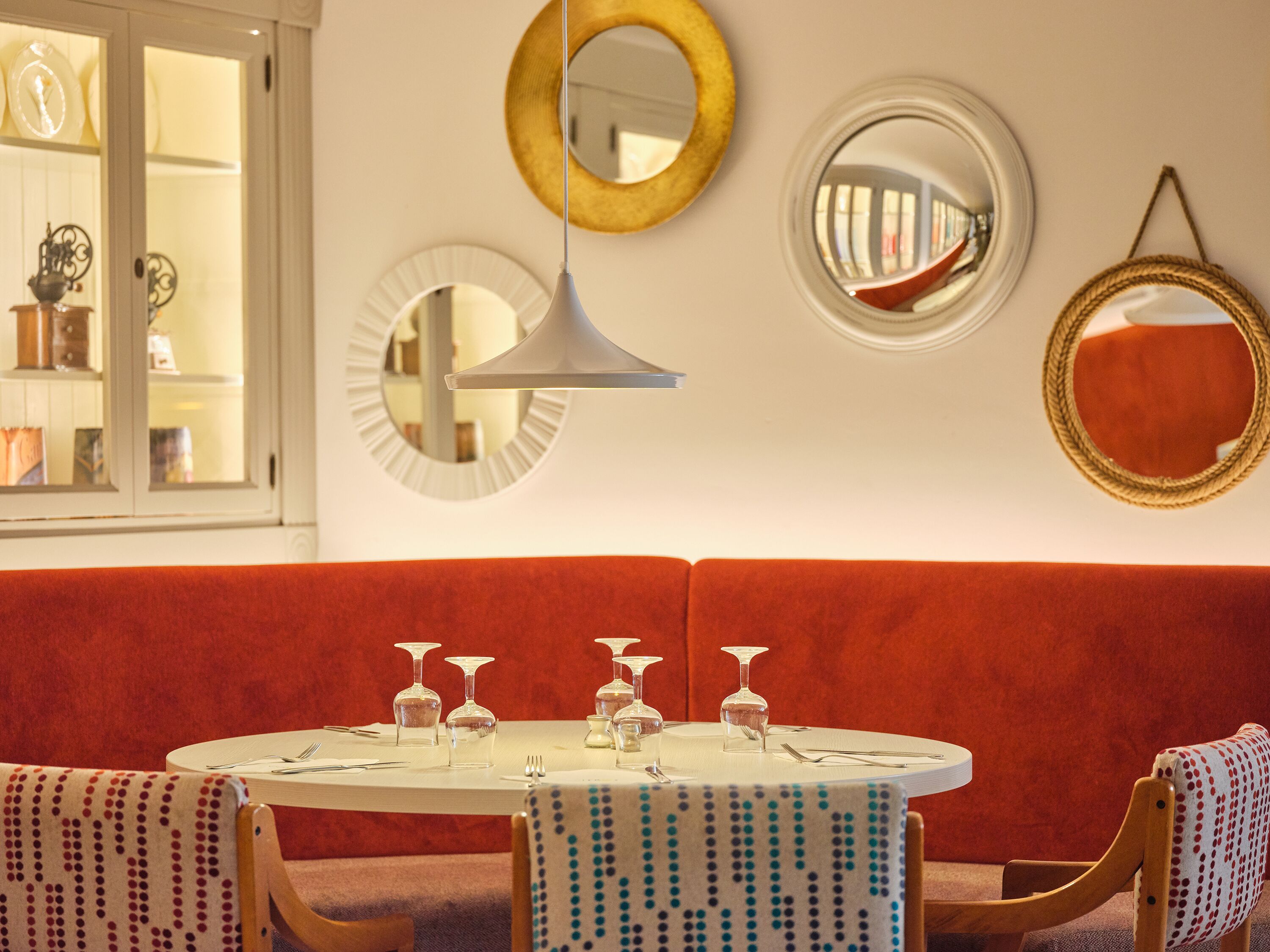 Un coin repas avec une table ronde dressée pour six personnes, entourée d’un canapé rouge recouvert de tissu et de chaises en bois.
