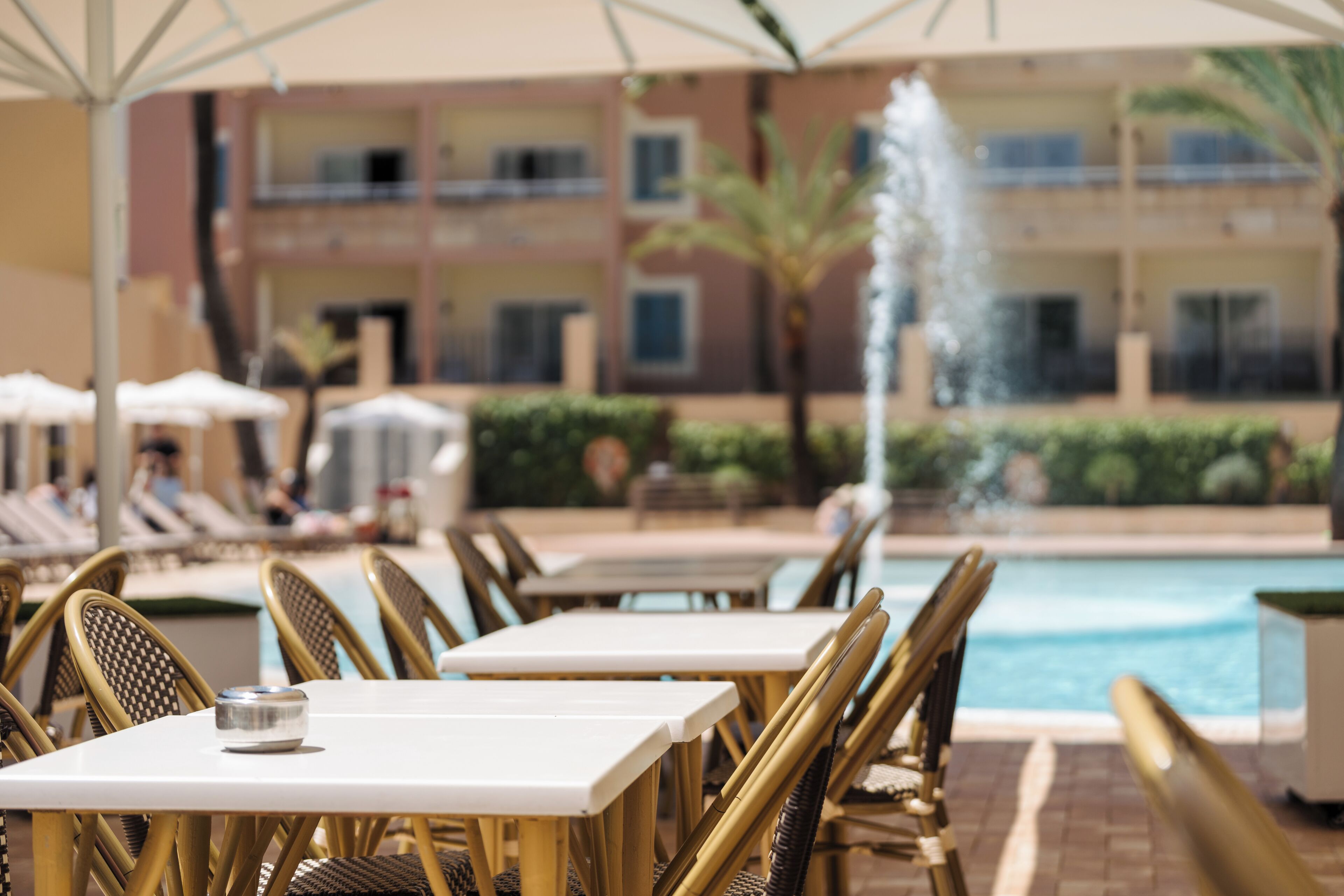 Un espace aménagé au bord de la piscine, avec des tables et des chaises, une fontaine, et un bâtiment doté de balcons en arrière-plan.