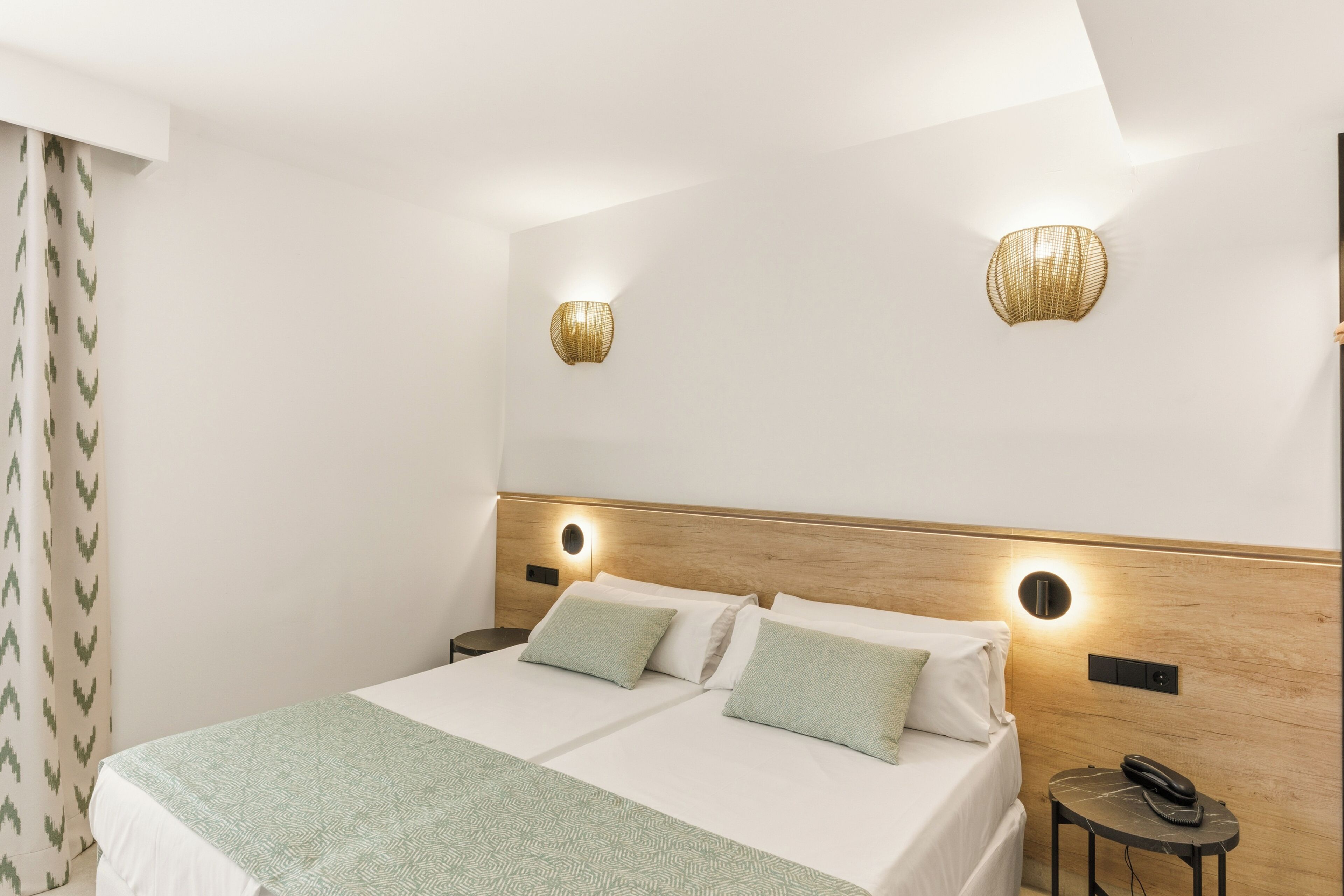 A hotel room with a bed, two bedside tables, a wall-mounted lamp, and a curtain.