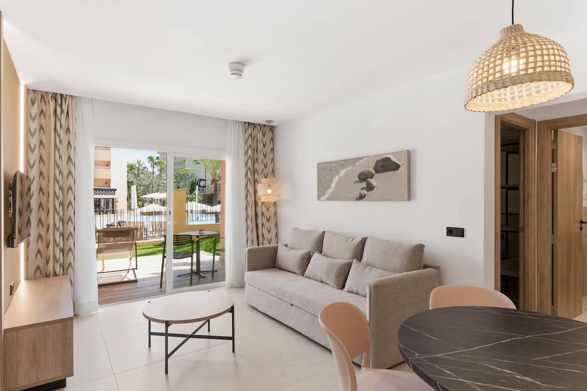 A modern living room with a sofa, dining table, and chairs, a hanging light fixture, and a view of the outdoor area.
