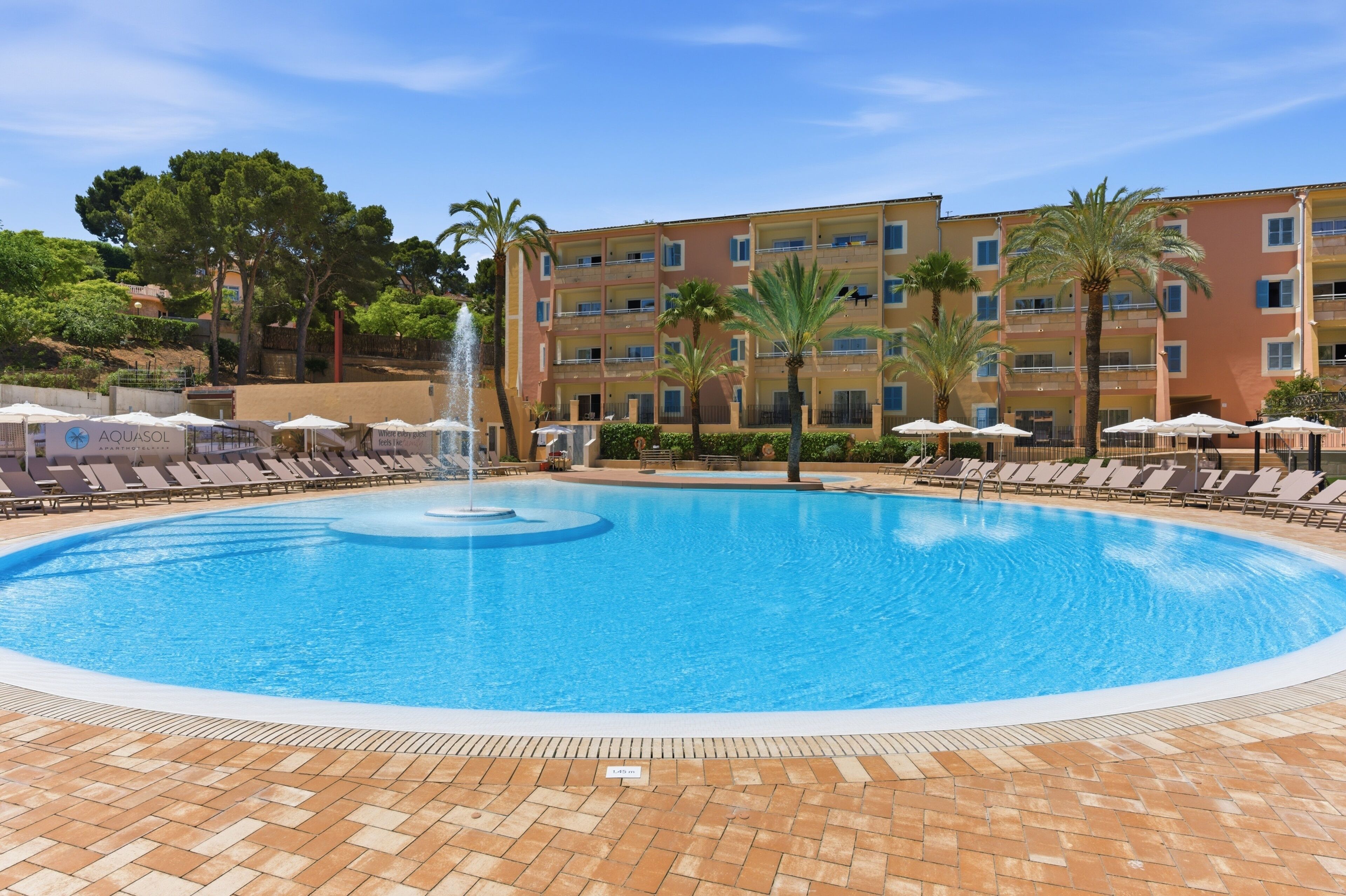 Una piscina con una fontana, circondata da lettini e ombrelloni, con un edificio a più piani sullo sfondo.