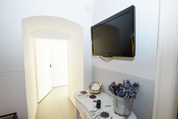 Una habitación con una televisión de pantalla plana en la pared, una mesa pequeña con un florero, una cesta y un control remoto.
