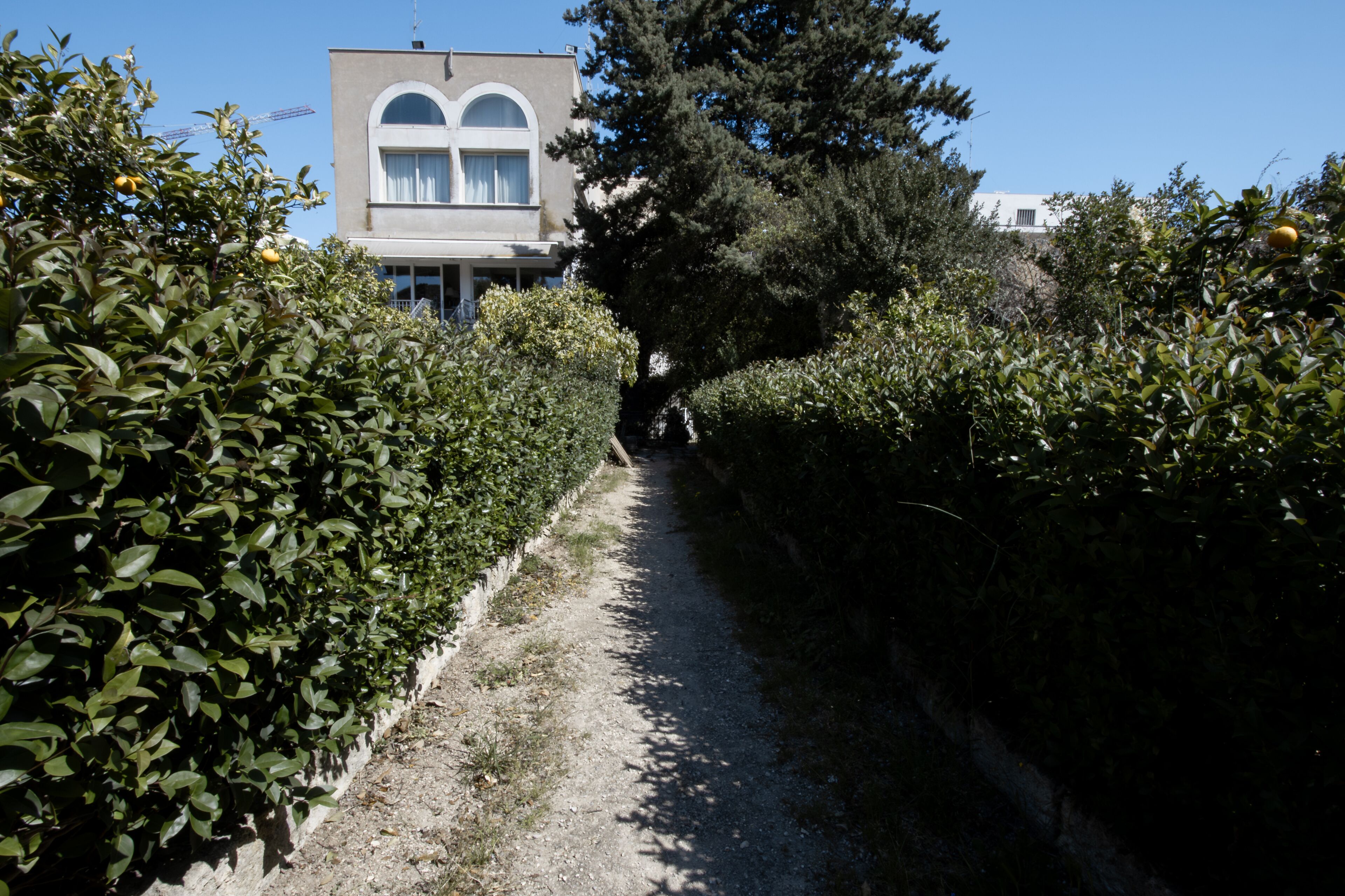 Un étroit sentier de gravier bordé d’une végétation luxuriante et verdoyante, qui mène à un bâtiment à la façade blanche et aux fenêtres en arc de cercle.