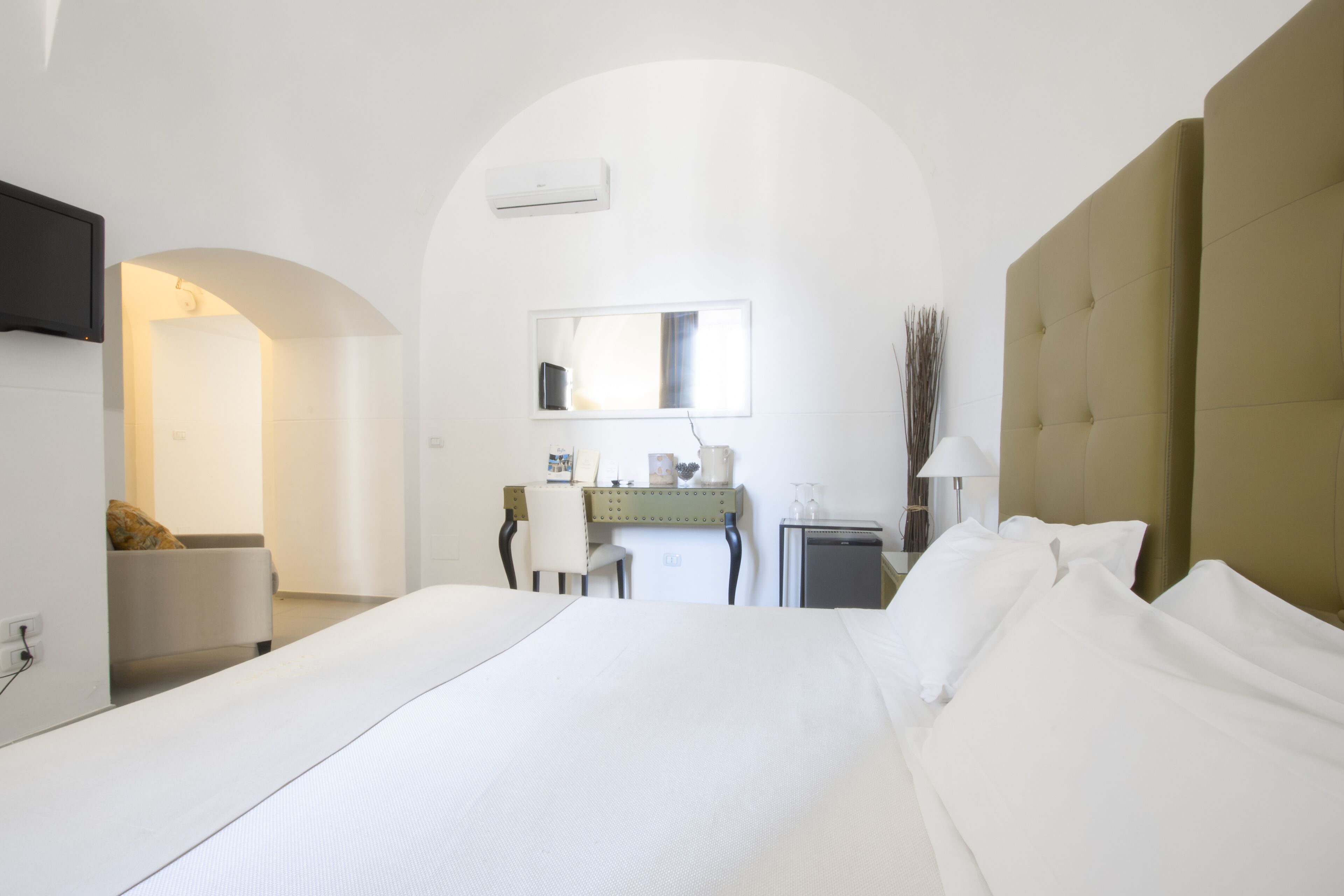 A modern hotel room with a large bed, a desk with a chair, a mirror, and an air conditioning unit.