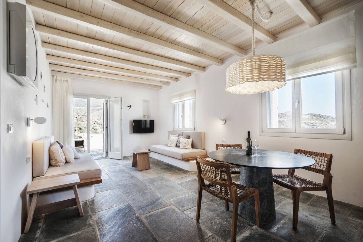 Un intérieur moderne avec un sol en pierre, des poutres en bois au plafond, une table à manger ronde entourée de chaises, un canapé et un téléviseur fixé au mur.
