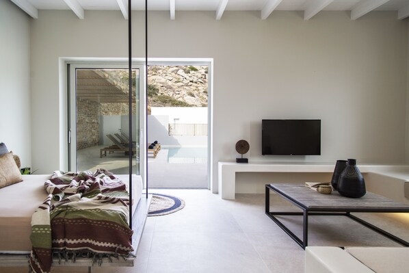 A modern living room with a sofa, a coffee table, and a flat-screen TV. There is a glass door leading to an outdoor area with a view of rocky terrain. - Hide out Suites (Ios)