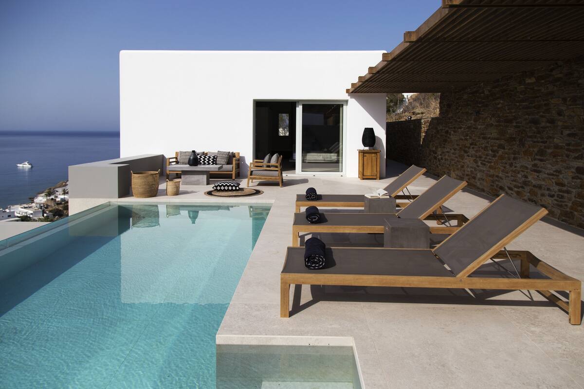 Une piscine à débordement avec des chaises longues, un mur en pierre et une vue sur la mer.