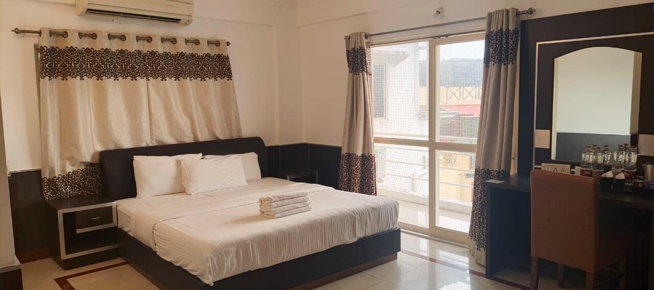 A hotel room with a bed, bedside tables, a desk with a chair, and a large window with curtains.