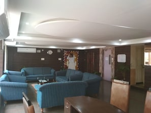 A hotel lobby with blue upholstered sofas arranged in a U-shape, a wooden reception desk, and a wall-mounted clock.